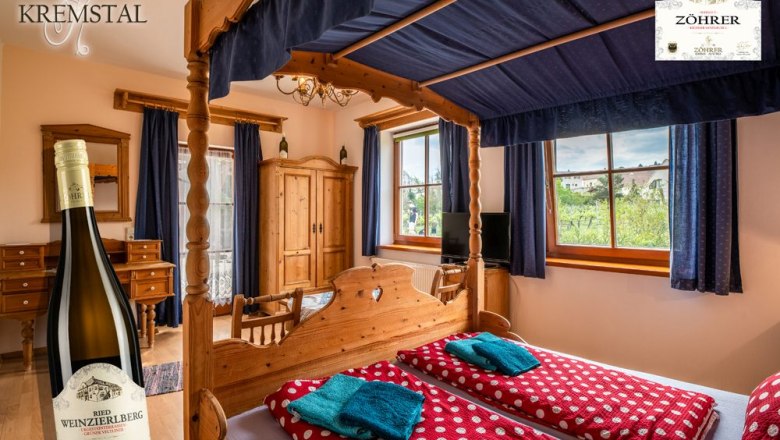 Zimmer Nr. 3 Himmelbett, © Weingut Zöhrer Ein gemütliches Zimmer mit Himmelbett, Holzmöbeln und Blick auf Weinberge. Eine Weinflasche ist im Vordergrund abgebildet.