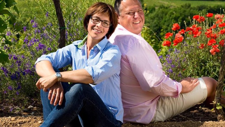 weingut-jschmid, © Weingut Josef Schmid / pov.at Zwei Personen sitzen Rücken an Rücken in einem Weinberg mit Blumen im Hintergrund.