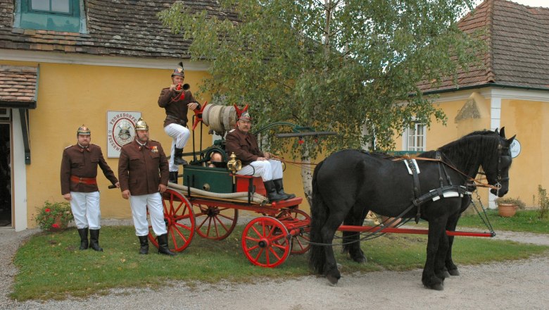 Feuerwehrmuseum Lanzenkirchen, © Gemeinde Lanzenkirchen Historische Feuerwehrkutsche mit Pferd und Feuerwehrleuten in Uniform vor einem gelben Gebäude.