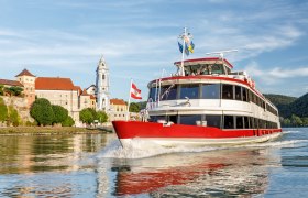 DDSG Blue Danube Schiffahrt, © DDSG Blue Danube Schiffahrt/Wöckinger Ein rotes Passagierschiff fährt auf der Donau vor einer historischen Stadt mit einem markanten Kirchturm.