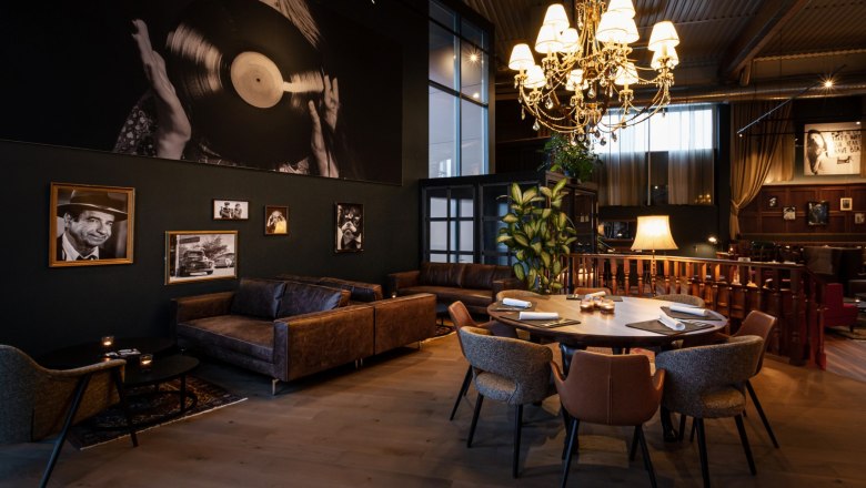 Zündwerk Living Room, © DERENKO DESIGN Ein stilvoll eingerichteter Raum mit Ledersofas, einem runden Tisch und einem großen Kronleuchter. An der Wand hängen Schwarz-Weiß-Fotografien.