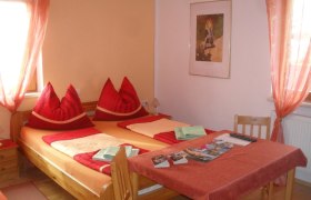 Schlafbereich beim Horak, © Hubert Horak Gemütliches Schlafzimmer mit Doppelbett, roten Kissen und Tisch mit Broschüren.