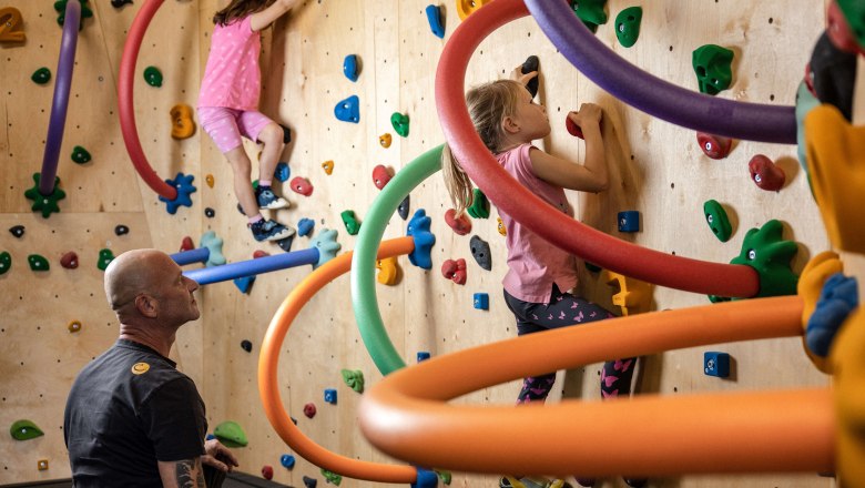 BigWall Bouldering Kinderkurs, © BigWall Bouldering Kinder klettern an einer bunten Kletterwand mit Schaumstoffringen, während ein Erwachsener beaufsichtigt.