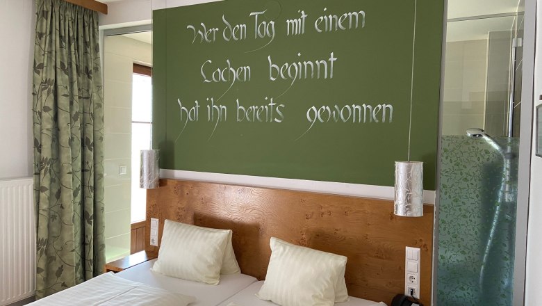 Doppelzimmer, © Wiener Alpen Ein Doppelzimmer mit zwei Betten, grüner Wand mit Spruch, Vorhängen und einer Dusche mit Glaswand.