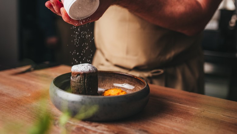 Schokolade und Mandarine, © Niederösterreich Werbung / Daniela Führer Person bestreut Schokoladenkuchen mit Puderzucker, daneben eine Mandarine auf einem Teller.
