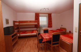 Gasthof Hödl - Tennishalle, © Gasthof Hödl Ein einfach eingerichtetes Zimmer mit Etagenbett, Einzelbett, Tisch und Stühlen. Ein Fernseher steht auf einem Regal. Fenster mit Vorhängen im Hintergrund.
