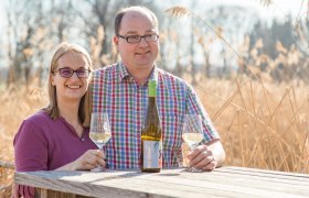 familyshooting-nolz-13_verena_saloukeh_1, © Verena Saloukeh Photography Ein Mann und eine Frau sitzen an einem Holztisch im Freien mit Weingläsern und einer Weinflasche.