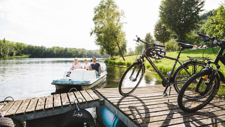 Tretbootfahren am Donausee in Weitenegg, © NÖW_Stefan Fürtbauer Drei Personen auf einem Tretboot auf einem See, daneben Fahrräder auf einem Steg.
