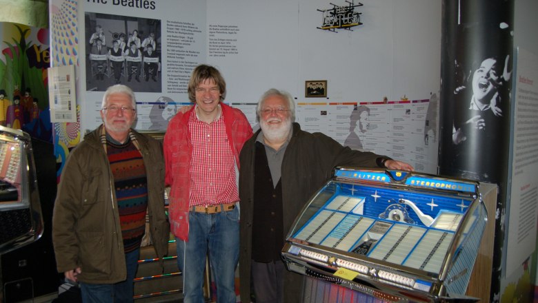 Pindigiland, © Pindigiland Drei Männer stehen vor einer Beatles-Ausstellung mit einer Jukebox.