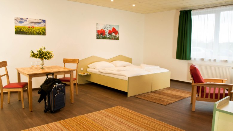 Studio, © Winrooms Betriebs GmbH Ein modernes Studio mit Doppelbett, Tisch und Stühlen, rotem Sessel und Koffer. Helle Wände, grüne Vorhänge und Blumenbilder an der Wand.