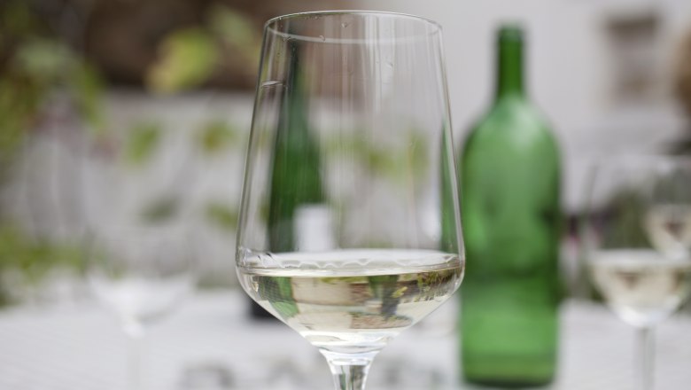 Weinglas, © Weinviertel Tourismus / Christine Wurnig Ein Weinglas mit Weißwein auf einem Tisch, im Hintergrund eine grüne Weinflasche.