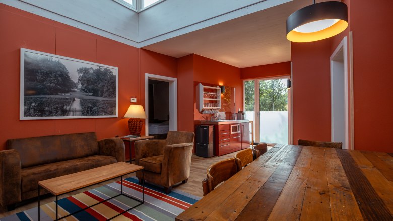 Grafenegg Cottages, © Niederösterreich Werbung / Maximilian Pawlikowsky Innenansicht eines modern eingerichteten Wohnzimmers mit roten Wänden, Holztisch, Sofa und Küchenzeile.