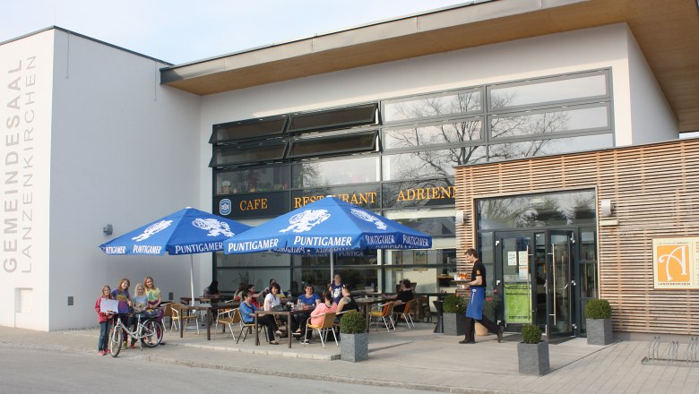 Cafe Adrienn, © Cafe Adrienn Außenansicht des Cafe Adrienn mit Gästen unter blauen Sonnenschirmen.