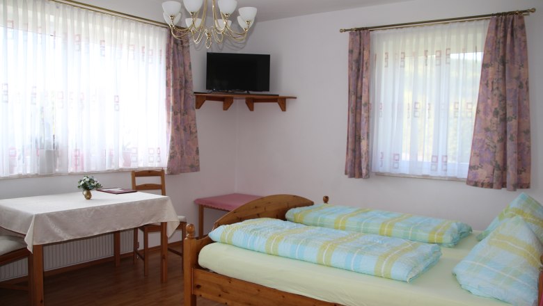 Mehrbettzimmer, © Karl Buchner Ein helles Zimmer mit Doppelbett, Tisch, Stühlen und einem Fernseher an der Wand.