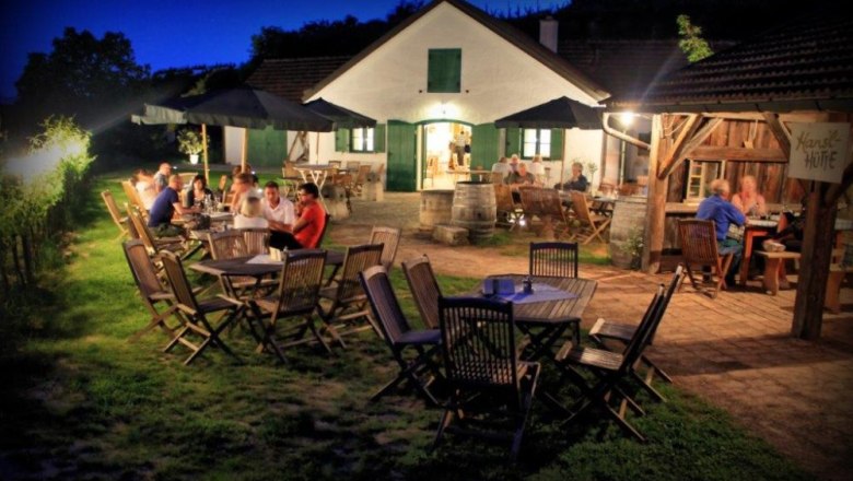 Abendstimmung beim Heurigen, © Johannes Kitzler Abendliche Szene in einem Heurigen mit Gästen an Holztischen im Freien.
