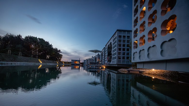 Therme Laa | Hotel | Silent Spa | Villas, © Niederösterreich Werbung / Maximilian Pawlikowsky Moderne Gebäude mit Wasserreflexionen bei Dämmerung.
