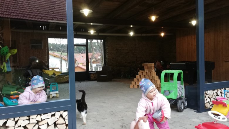 Streicheltierstall, © Familie Rosinger Ein Kind in Winterkleidung spielt mit einem Spielzeugpferd in einem offenen Raum mit Holzstapeln und Spielzeug. Eine Katze läuft vorbei.