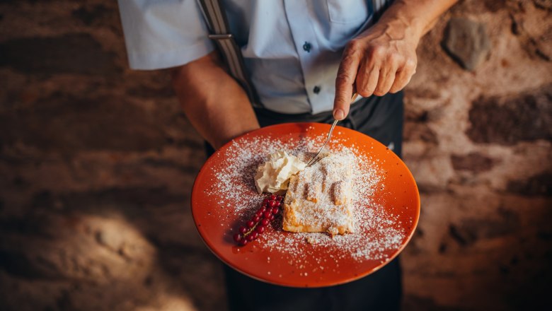 Apfelstrudel, © Niederösterreich Werbung/Daniela Führer Ein Teller mit Apfelstrudel, Sahne und Johannisbeeren, gehalten von einer Person in einem blauen Hemd.