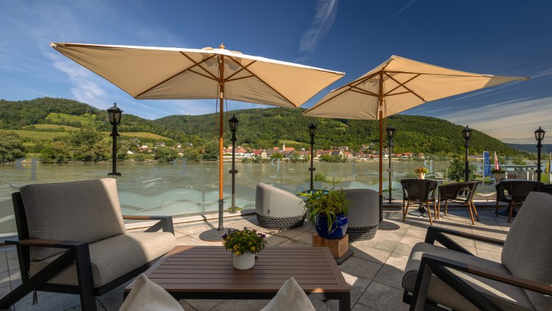 Hotel ResidenzWachau, © Niederösterreich Werbung / Maximilian Pawlikowsky Terrasse mit Blick auf die Donau und umliegende Hügel, ausgestattet mit Sonnenschirmen und Sitzmöbeln.