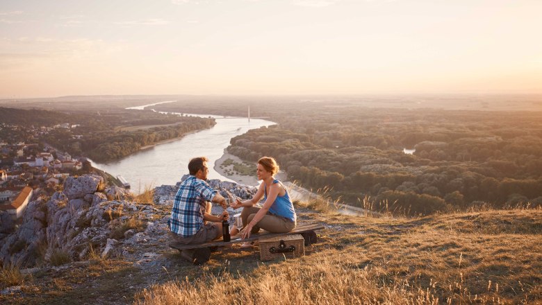 Blick vom Braunsberg, Hainburg/Donau, © Donau Niederösterreich, Andreas Hofer Zwei Personen sitzen auf einem Hügel mit Blick auf einen Fluss und eine Stadt im Sonnenuntergang.