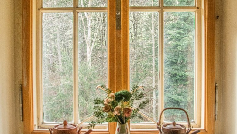 Orlet an der Rax, © Sofia Orlet Fenster mit zwei Teekannen und einer Vase mit Blumen auf der Fensterbank, Wald im Hintergrund.