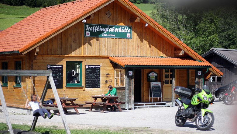 Trefflingtalerhütte, © weinfranz.at Eine Holzhütte mit dem Schild 'Trefflingtalerhütte', davor eine Schaukel, ein Motorrad und ein Mensch an einem Tisch.