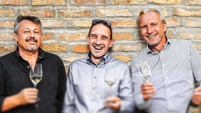Weingut Dürnberg, © Weingut Dürnberg Drei Männer stehen vor einer Ziegelwand und halten Weingläser in der Hand.