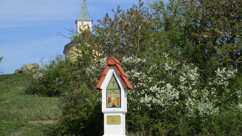 Michelberg, © Karoline Krammer Eine kleine Kapelle mit grünem Turm hinter blühenden Büschen und einem Bildstock im Vordergrund.
