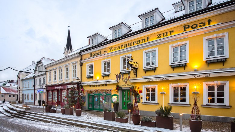 Aussenansicht im Winter Hotel-Restaurant Zur Post, © Helmut Lackinger Außenansicht eines gelben Gebäudes mit der Aufschrift 'Hotel-Restaurant zur Post' im Winter, umgeben von Schnee und dekoriert mit Lichtern.