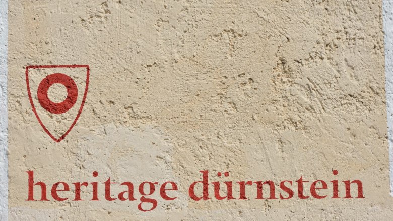Heritage Dürnstein, © Gregor Semrad Wand mit rotem Schriftzug 'heritage dürnstein' und einem roten Symbol.