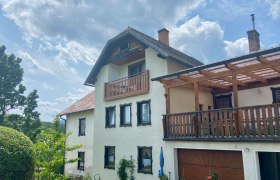 Ferienwohnungen Riedl, © Wiener Alpen Ein zweistöckiges Haus mit Balkon und Terrasse, umgeben von Garten und Bäumen, unter einem bewölkten Himmel.