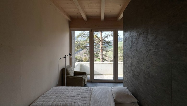 Schlafbereich, © Meinrad Hofer Minimalistisches Schlafzimmer mit Bett, Sessel und Fenster mit Blick auf Bäume.