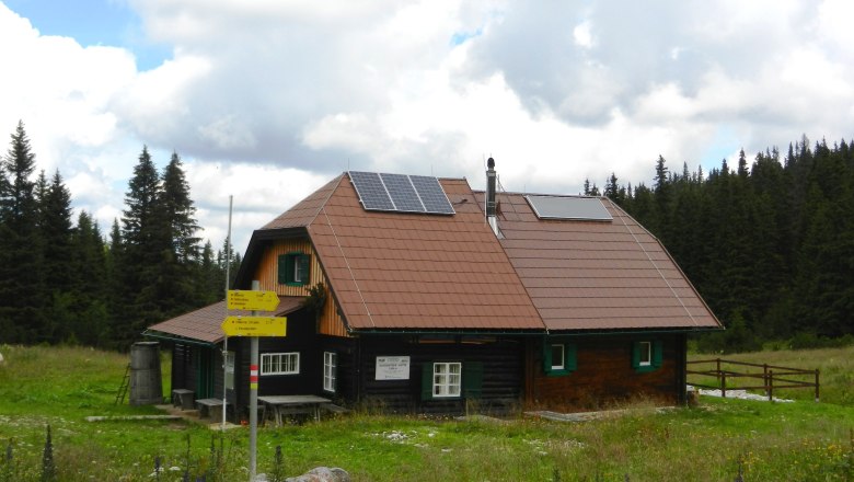 Huette3, © Richard Waidhofer Eine Berghütte mit Solarpanelen auf dem Dach, umgeben von Wiesen und Wald.