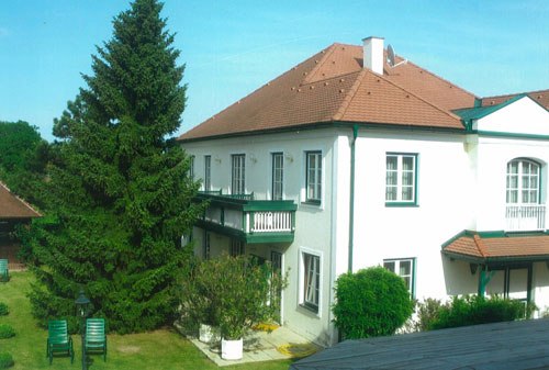 Gästehaus, © Fam. Weber Ein zweistöckiges weißes Gästehaus mit rotem Dach und grünen Fensterläden, umgeben von einem Garten mit Bäumen und Gartenmöbeln.