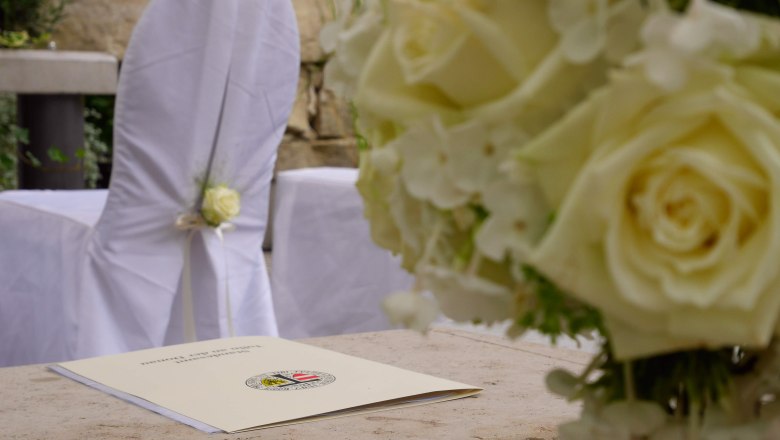 Hochzeit im Liebesgarten, DIE GARTEN TULLN, © DIE GARTEN TULLN Nahaufnahme einer Hochzeitsdekoration mit weißen Rosen und einem Dokument auf einem Tisch.