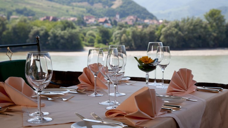 Restaurant-Terrasse, © Fally Gedeckter Tisch auf einer Restaurant-Terrasse mit Blick auf einen Fluss und bewaldete Hügel im Hintergrund.