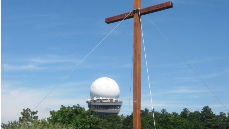 Buschberg, © Gemeinde Niederleis Ein Holzkreuz vor einer Radarkuppel auf dem Buschberg, umgeben von Bäumen und blauem Himmel.