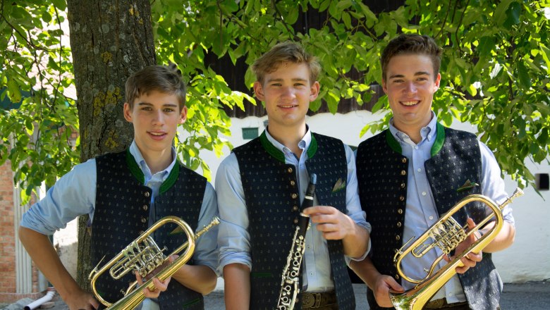 Honsig Buam, © Thomas Honsig Drei junge Männer in traditioneller Kleidung mit Blasinstrumenten vor einem Baum.