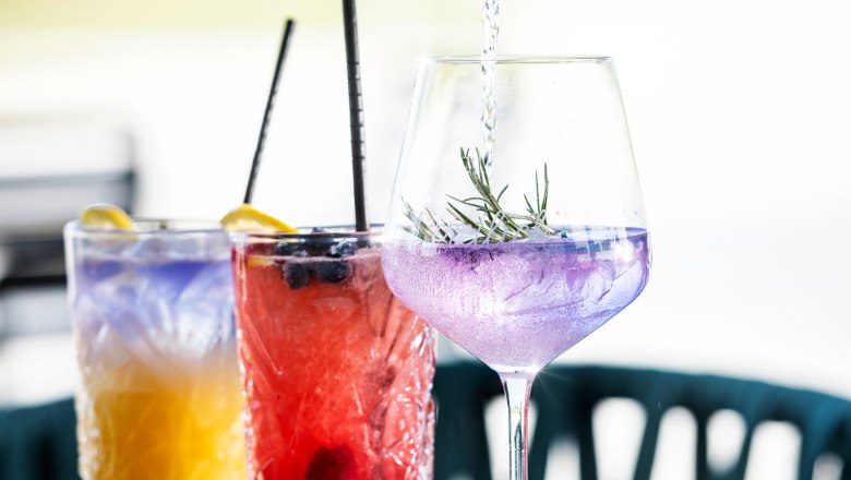 Cocktails, © Alexander Künzl Photography Drei bunte Cocktails auf einem Tisch, eine Hand gießt Flüssigkeit in ein Glas.