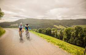Rennradfahren bei Kirchschlag in der Buckligen Welt, © Wiener Alpen, Martin Fülöp Zwei Radfahrende auf einer kurvigen Straße in einer hügeligen Landschaft mit der Burgruine Kirchschlag im Hintergrund.