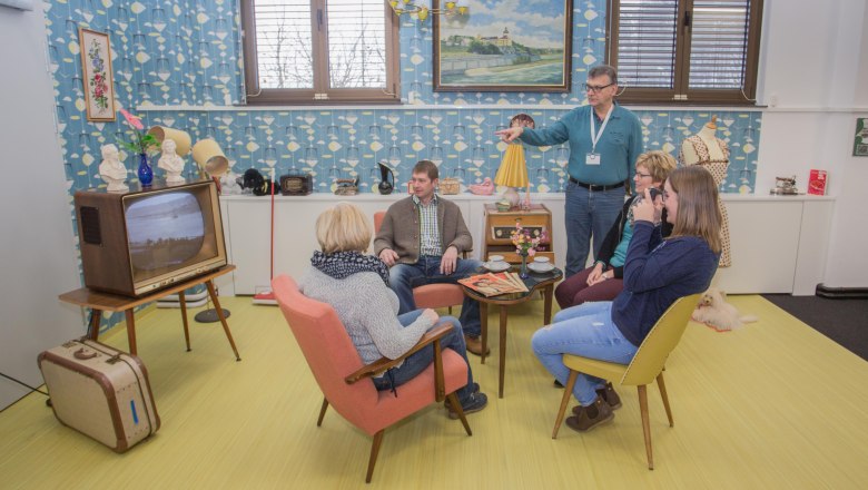 60er-Jahre-Wohnzimmer, © Franz Engl Gruppe von Menschen in einem Wohnzimmer im Stil der 60er Jahre mit Retro-Möbeln und Dekoration.