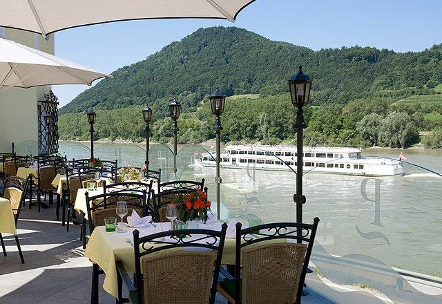 Terrasse, © Residenz Wachau Terrasse mit gedeckten Tischen, Blick auf Fluss und vorbeifahrendes Schiff, umgeben von grünen Hügeln.