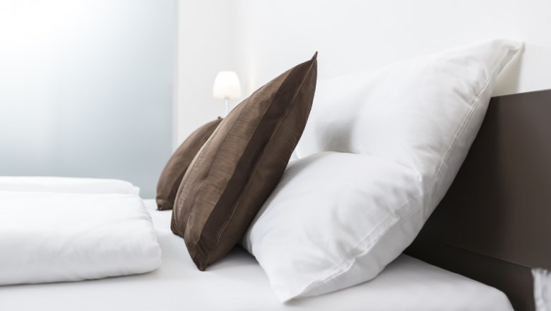 Gemütliches Bett, © Ernst Jöchlinger Ein Bett mit weißen Kissen und braunen Zierkissen, im Hintergrund eine Lampe.