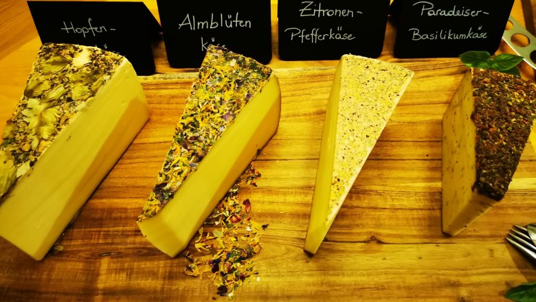 Affinierte Kaesespezialitaeten, © Michi's Käseladen Vier Käsesorten auf einem Holzbrett, beschriftet mit Hopfen-, Almblüten-, Zitronen-Pfeffer- und Paradeiser-Basilikumkäse.