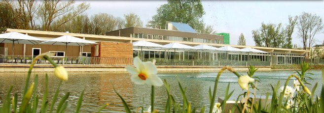 Blick auf Teich, © Josef Pichler Ein modernes Gebäude mit Terrasse und Sonnenschirmen an einem Teich mit Blumen im Vordergrund.