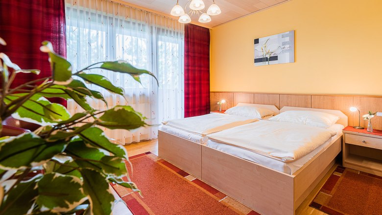 zimmer01, © Hotel Garni Steinfeld Gemütliches Schlafzimmer mit Doppelbett, gelber Wand, rotem Teppich und Pflanzen im Vordergrund.