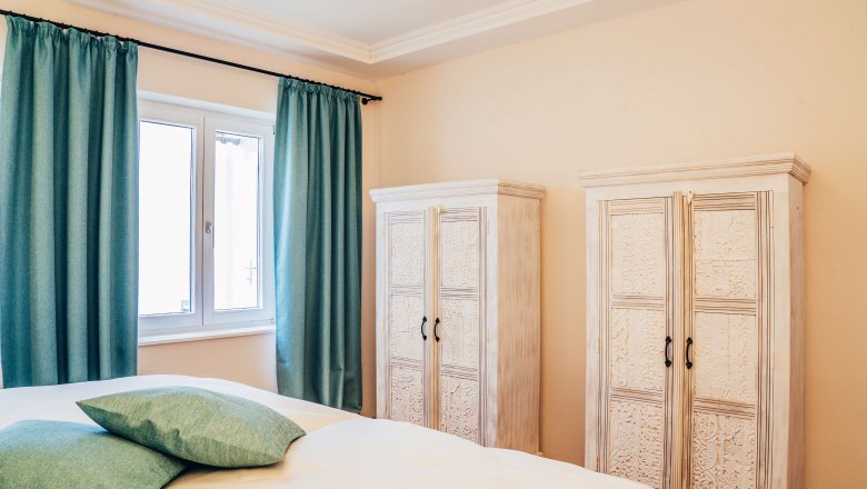 Roth0_4, © Georg Pomassl Schlafzimmer mit zwei weißen Schränken, türkisfarbenen Vorhängen und Kissen.