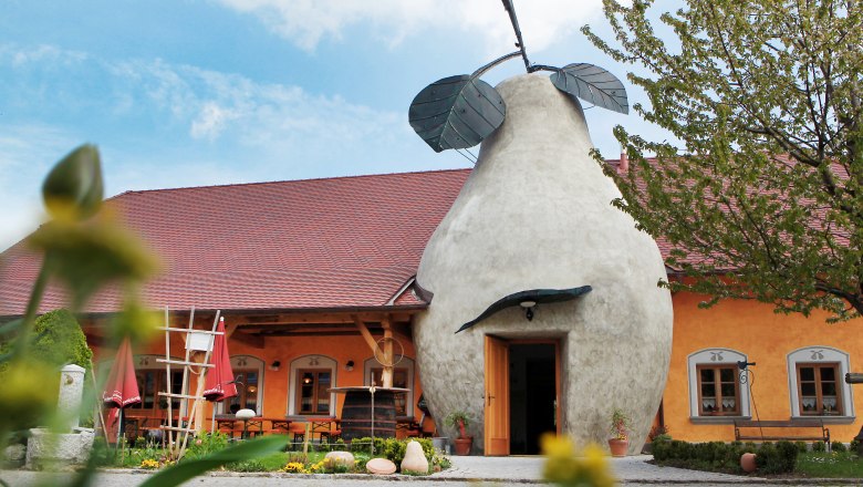 Zur steinernen Birne, © weinfranz.at Gebäude mit birnenförmigem Eingang und rotem Dach, umgeben von Pflanzen.