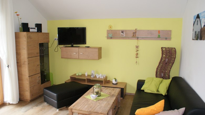 Wohnraum, © Familie Zwinz Modernes Wohnzimmer mit gelber Wand, schwarzem Sofa, Holzmöbeln und Dekorationen.