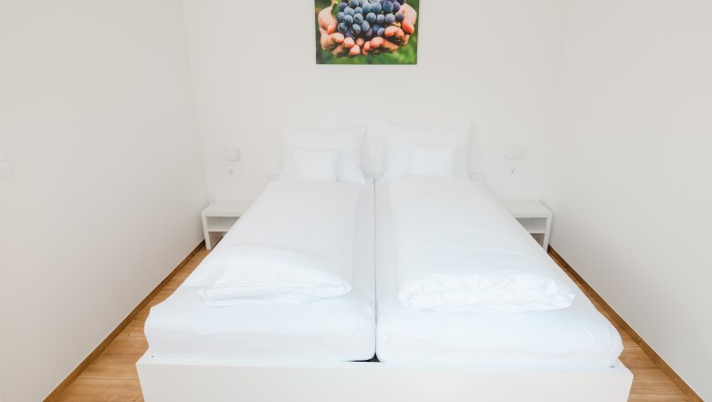 Ipsmiller Appartements, © Michael Reidinger Ein schlichtes, modernes Schlafzimmer mit einem Doppelbett und einem Bild von Trauben an der Wand.